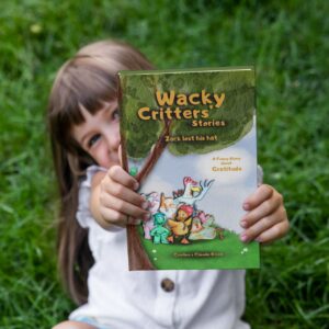 A little smiling girl, sitting on a green meadow, holding in her hands the hardcover copy and our first children's story: Wacky Critters' Stories, Zack lost his hat