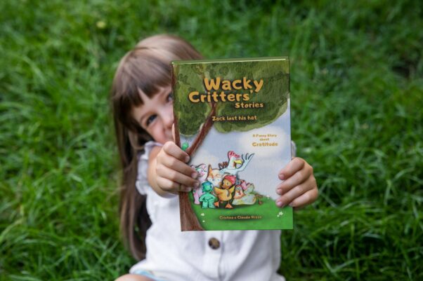 A little smiling girl, sitting on a green meadow, holding in her hands the hardcover copy and our first children's story: Wacky Critters' Stories, Zack lost his hat