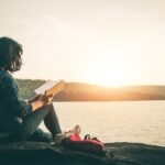 Young girl reading a self help book, sitting on a rock near the sea, while the sun sets