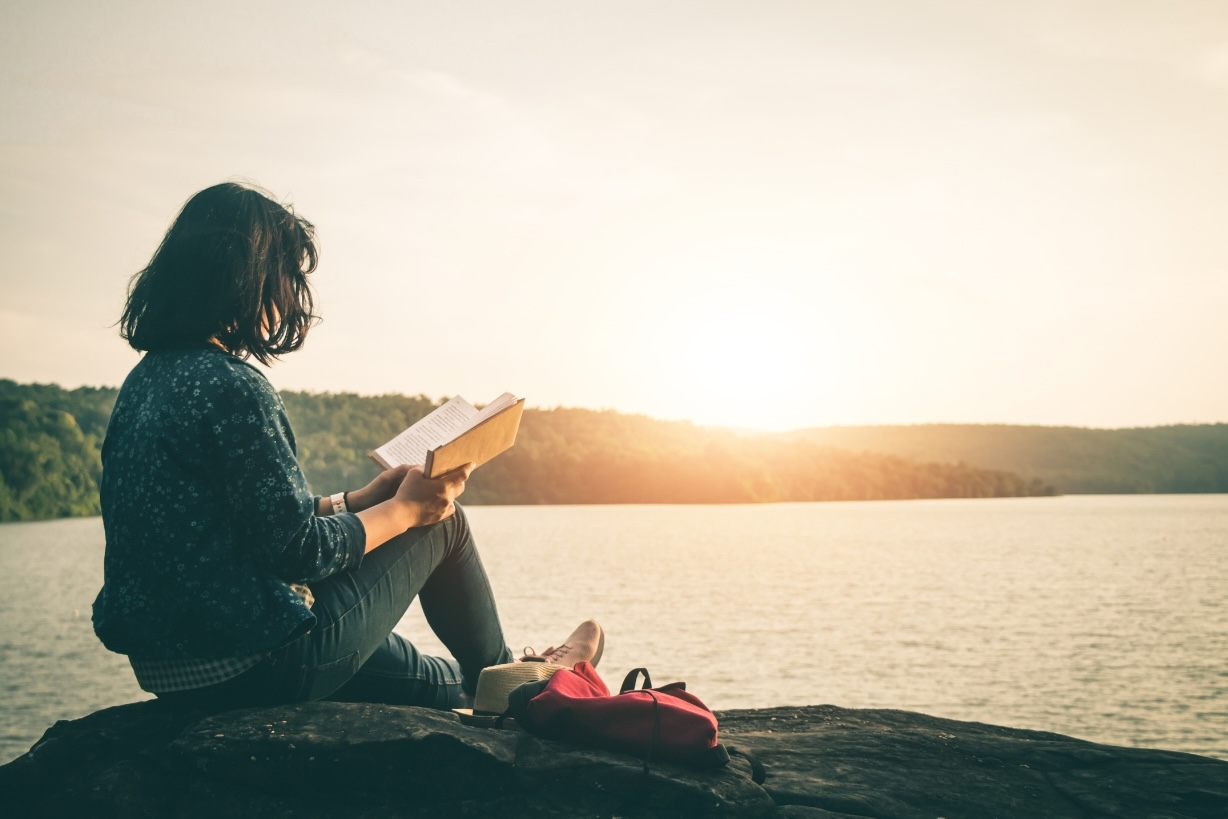 Young girl reading a self help book, sitting on a rock near the sea, while the sun sets